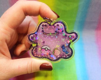 May include: A purple and pink glittery charm shaped like a Pokemon character, with a clear resin background. The charm features a small Pokeball, a peach, and a candy cane.
