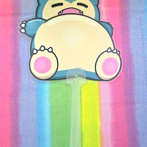 May include: A hand fan with a Snorlax cartoon design. The fan has a light blue border, a beige body, and pink accents on the feet. The handle is a translucent green colour. The background is a rainbow of colours.