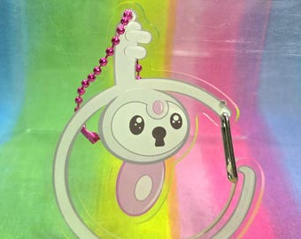 Puede incluir: Un llavero acrílico transparente con un personaje de dibujos animados con cuerpo blanco, detalles rosas y diseño de ojo de cerradura. El llavero tiene un cierre plateado y una cadena de cuentas rosas. El fondo es un degradado arcoíris.