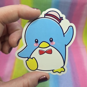 Pode incluir: Um autocolante com um pinguim de desenho animado com corpo azul, barriga branca e pés amarelos. O pinguim usa uma gravata borboleta vermelha e um chapéu com uma faixa vermelha. O autocolante tem uma borda branca e é segurado por uma mão.