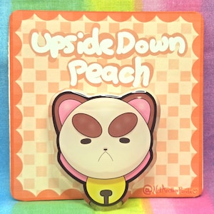 May include: A cartoon-style pin featuring a bear-like character with a grumpy expression. The pin is set against a square background with the words "Upside Down Peach" in a playful font. The background is orange with a checkered pattern.