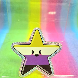 Könnte beinhalten: Ein sternförmiger Aufkleber mit dem Design der Non-Binary-Pride-Flagge. Der Aufkleber hat eine gelbe Oberseite, eine weiße Mitte, einen lila Boden und eine schwarze Basis. Der Stern hat einen glitzernden silbernen Rand und ein niedliches Gesicht.