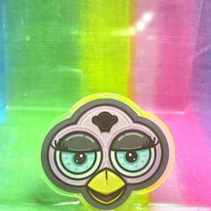 May include: A colorful sticker featuring a stylized Furby face. The design includes large, blue eyes with long eyelashes, a yellow beak, and a pink and black outline. The sticker has a glossy finish and is set against a rainbow background.