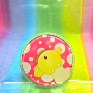 May include: A circular pin badge with a cartoon design. It features a yellow character with wings and a light beige character, set against a pink background with white polka dots. The badge is outlined in green.