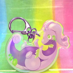 Peut inclure: Un porte-clés en acrylique transparent représentant un dragon de dessin animé violet avec des accents verts. Le dragon est enroulé sur lui-même, avec un fermoir argenté en haut. Le porte-clés a un effet holographique, avec un fond arc-en-ciel.