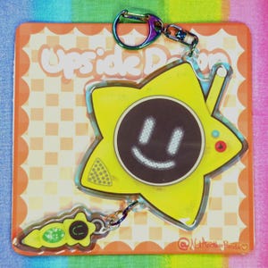 May include: A yellow star-shaped keychain with a smiling face on a black circle. The keychain has a rainbow-colored clasp and a smaller, green and yellow charm. The background features the text "Upside Down" and a checkered pattern.