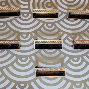 May include: Seven rectangular cabinet pulls with a black center and a decorative copper-colored top and bottom edge. The pulls are arranged on a patterned background with a gold and white design.