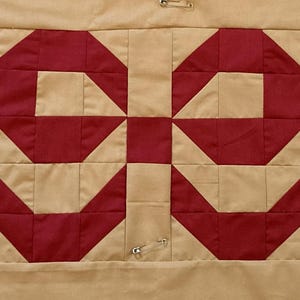 Op de afbeelding: Close-up van een quilt met een herhalend geometrisch patroon. Het ontwerp heeft een symmetrische opstelling van rode vormen tegen een beige achtergrond. Veiligheidsspelden zijn zichtbaar, die de lagen bij elkaar houden. De quilt is waarschijnlijk handgemaakt.