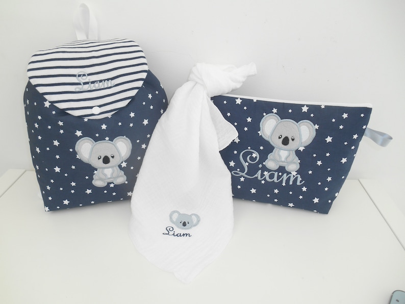K&ouml;nnte beinhalten: Ein blaues und wei&szlig;es Wickeltaschen-Set mit Koala-Motiv. Das Set beinhaltet einen Rucksack, ein Waschlappen und eine Kosmetiktasche. Alle Artikel sind mit dem Namen "Liam" bestickt.