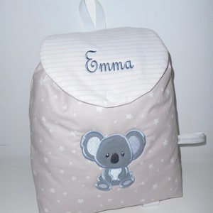 K&ouml;nnte beinhalten: Ein beiger Kinderrucksack mit einem wei&szlig;en Sternenmuster. Der Rucksack hat eine Klappe mit einem wei&szlig;en und rosa gestreiften Muster und den Namen "Emma" darauf gestickt. Ein grauer Koalab&auml;r ist auf der Vorderseite des Rucksacks gestickt.
