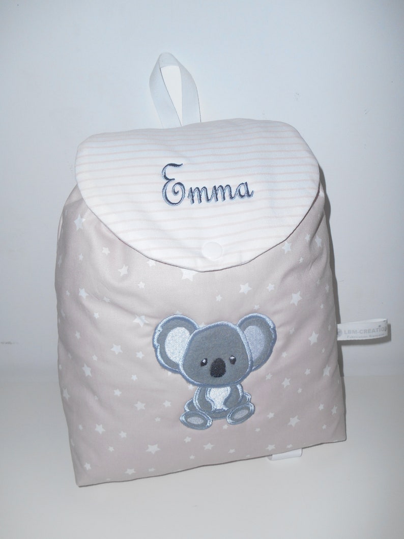 K&ouml;nnte beinhalten: Ein beiger Kinderrucksack mit einem wei&szlig;en Sternenmuster. Der Rucksack hat eine Klappe mit einem wei&szlig;en und rosa gestreiften Muster und den Namen "Emma" darauf gestickt. Ein grauer Koalab&auml;r ist auf der Vorderseite des Rucksacks gestickt.