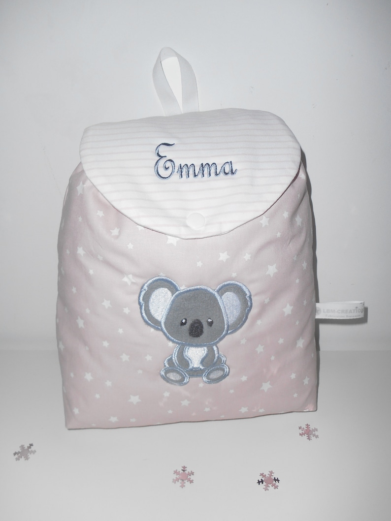 K&ouml;nnte beinhalten: Ein rosa Stoffrucksack mit wei&szlig;en Sternen und einem gestickten grauen Koala auf der Vorderseite. Der Rucksack hat eine wei&szlig;e Klappe mit rosa Streifen und den Namen "Emma" darauf gestickt.