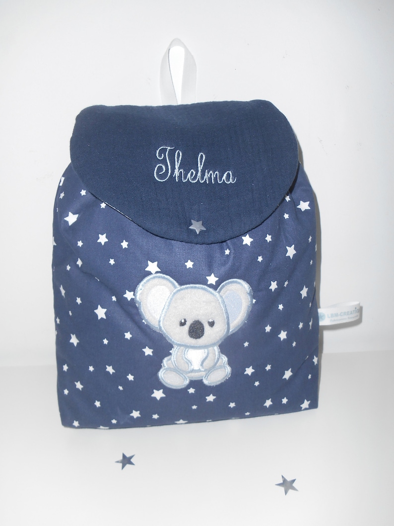 K&ouml;nnte beinhalten: Ein blauer Rucksack mit wei&szlig;en Sternen und einem gestickten grauen Koala auf der Vorderseite. Der Rucksack hat eine Klappe mit dem gestickten Namen "Thelma".