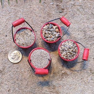 May include: Four miniature red buckets filled with different types of sand and gravel. The buckets are arranged in a circle on a light brown sandy surface.