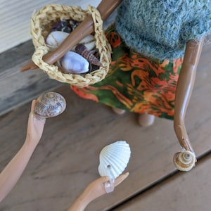 May include: A doll with brown hair and a blue and green patterned dress holds a woven basket filled with seashells. The doll is holding a seashell in one hand and another seashell is on the floor.