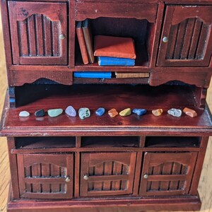 May include: A miniature wooden hutch with two doors on top and three doors on the bottom. The top shelf has books and the bottom shelf has a row of small colorful stones.