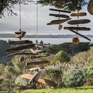 May include: Driftwood wind chimes suspended from a tree branch, composed of natural wood pieces, stones, and shells. The chimes are hung by twine, creating a rustic, organic aesthetic. The background shows a scenic view of a body of water and lush greenery.