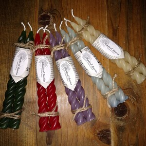 May include: Six twisted candles in various colors, including green, red, purple, and cream, are bundled together with twine. Each candle is labeled with a white sticker that reads "Mystical Sunrise Creative Candles".