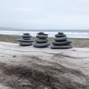 May include: Three stacks of grey and black stones balanced on a weathered driftwood log. The stones are arranged in a pyramid shape, with the largest stone at the bottom and the smallest stone at the top. The background is a blurry image of a beach with waves crashing on the shore.