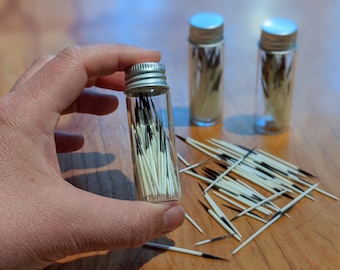 May include: Three small glass jars with silver lids filled with white and black feathers. The jars are on a wooden surface and some feathers are scattered around them.
