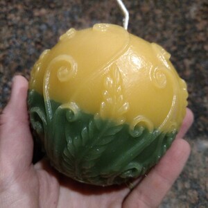May include: A round, yellow and green candle with a white wick. The candle has a floral design.