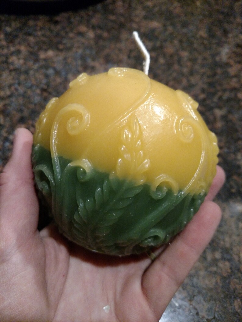 May include: A round, yellow and green candle with a white wick. The candle has a floral design.