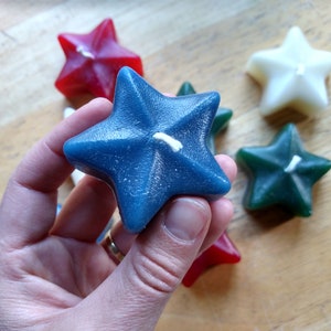 May include: A hand holds a blue star-shaped candle with a white wick. The candle is surrounded by other star-shaped candles in red, white, and green.