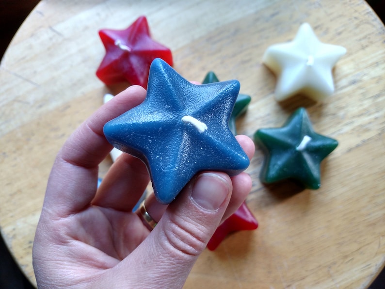 May include: A hand holds a blue star-shaped candle with a white wick. The candle is surrounded by other star-shaped candles in red, white, and green.
