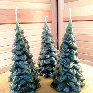 May include: Three green pine tree shaped candles. The candles are on a wooden surface.