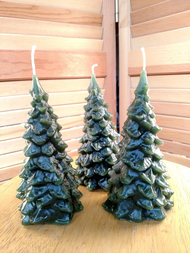 May include: Three green pine tree shaped candles. The candles are on a wooden surface.