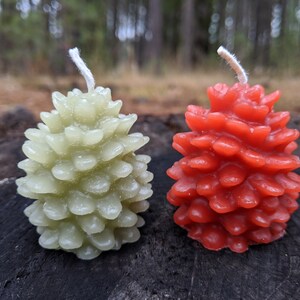 May include: Two pine cone shaped candles, one green and one red, on a wooden surface.