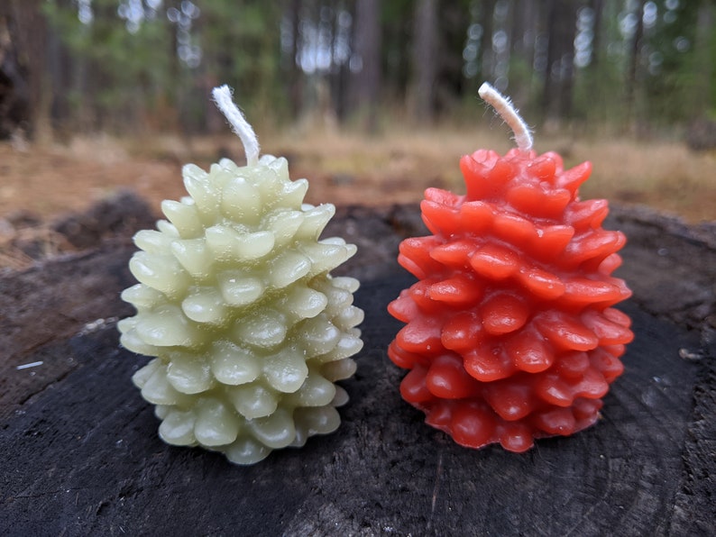 May include: Two pine cone shaped candles, one green and one red, on a wooden surface.