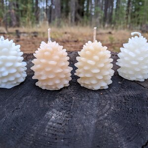 May include: Four pine cone shaped candles in white, cream, and light brown. The candles are arranged in a row on a dark brown wooden surface.