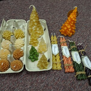 May include: Assortment of beeswax candles in various shapes and sizes, including pine tree and honeycomb designs. The candles are arranged in a cardboard egg carton and on a table. Some candles are tied with twine.