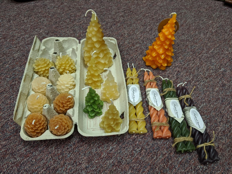 May include: Assortment of beeswax candles in various shapes and sizes, including pine tree and honeycomb designs. The candles are arranged in a cardboard egg carton and on a table. Some candles are tied with twine.