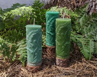 Puede incluir: Tres velas verdes de pilar con diseños de helechos. Las velas están sobre un lecho de agujas de pino.