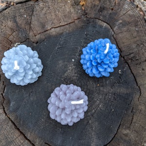 May include: Three pine cone shaped candles in shades of blue and gray. The candles are arranged on a wooden surface.