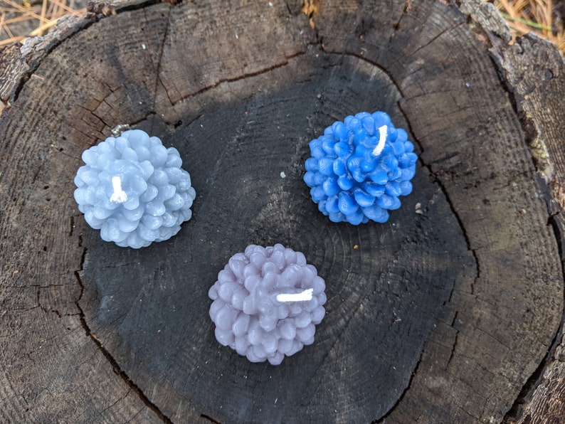 May include: Three pine cone shaped candles in shades of blue and gray. The candles are arranged on a wooden surface.