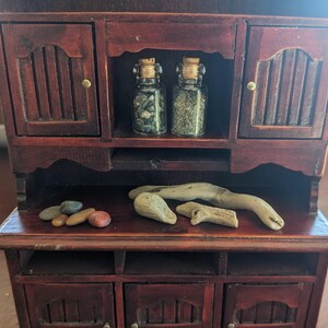May include: A miniature wooden cabinet with a dark brown finish. Two small glass jars with cork stoppers sit inside, filled with small stones. Several smooth stones and pieces of driftwood are arranged on the cabinet's surface.