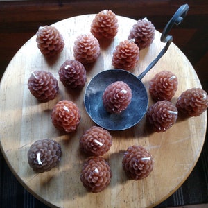 May include: A collection of brown pine cone shaped candles arranged on a wooden surface. The candles are all lit and have a warm glow.