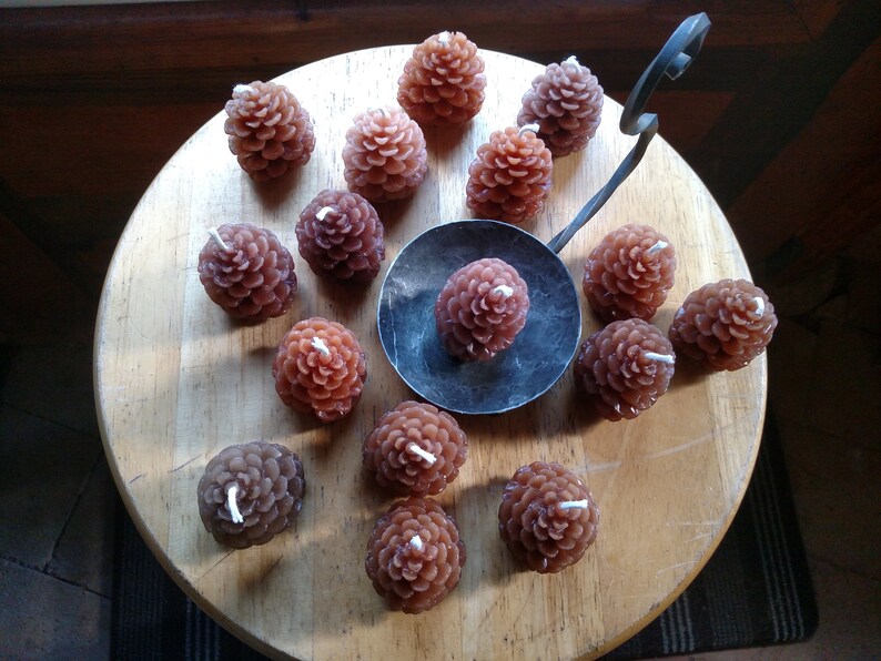 May include: A collection of brown pine cone shaped candles arranged on a wooden surface. The candles are all lit and have a warm glow.