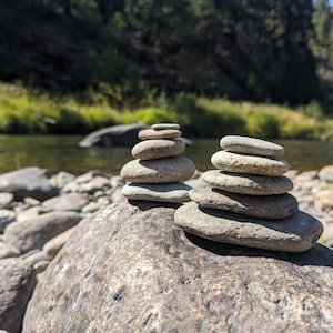 May include: Two stacks of smooth, grey river stones balanced on a large rock. The stones are various sizes and shapes, creating a sense of balance and tranquility. The background features a river and green foliage.