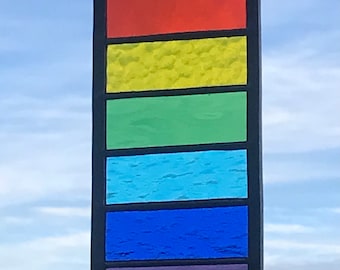 Könnte beinhalten: Ein Buntglasfenster mit einem Regenbogenmuster. Das Fenster besteht aus sieben horizontalen Glasbändern in den Farben Rot, Orange, Gelb, Grün, Blau, Hellblau und Lila.