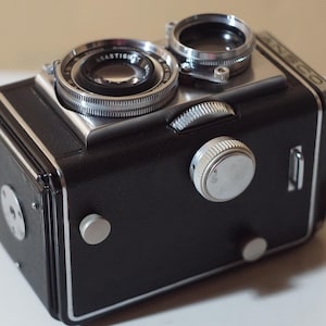 Pode incluir: Uma câmara reflex de objetiva dupla preta com detalhes em metal prateado. A câmara apresenta duas lentes circulares na parte frontal superior, um grande botão de focagem prateado na lateral e a palavra ANECO em letras prateadas no canto superior.