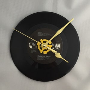 May include: A black vinyl record clock with gold hands. The record label features the song "Luka" by Suzanne Vega and the text "AM Records".