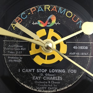 May include: A black vinyl record clock with gold-colored hands and a yellow center. The record label reads "I Can't Stop Loving You" by Ray Charles, with "ABC-Paramount" branding. The clock hands point to approximately 10:10.