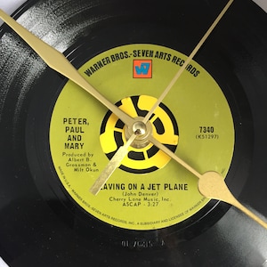May include: A black vinyl record clock featuring a yellow and black label with the text "Peter, Paul and Mary" and "Leaving on a Jet Plane (John Denver)". The record is labeled "Warner Bros.-Seven Arts Records" and has the number "7340 (K51297)" printed on it.