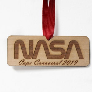 Peut inclure: Une décoration en bois avec un ruban rouge. La décoration est gravée avec le logo de la NASA et le texte "Cape Canaveral 2019".