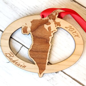 May include: A wooden ornament shaped like the country of Bahrain with the year 2017 and the word "Bahrain" engraved on it. The ornament is attached to a red ribbon.