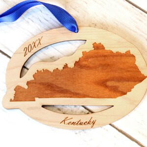 May include: A wooden ornament shaped like the state of Kentucky with the year 20XX and the word "Kentucky" engraved on it. The ornament is attached to a blue ribbon.